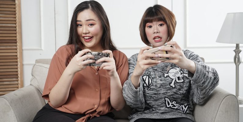 two women holding controllers sitting on sofa chair viral video, gaming audience, laughing viewers