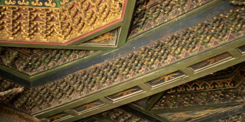 Intricately painted ornate ceiling with detailed patterns. ancient manuscript palm leaf, meditation posture, temple interior